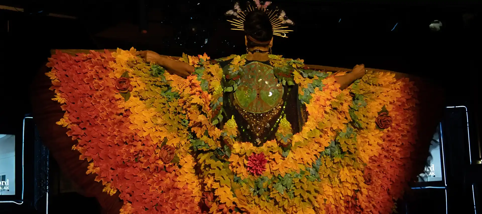 A woman wearing a coat made with individually colored leaves that appear like wings as she speads her arms